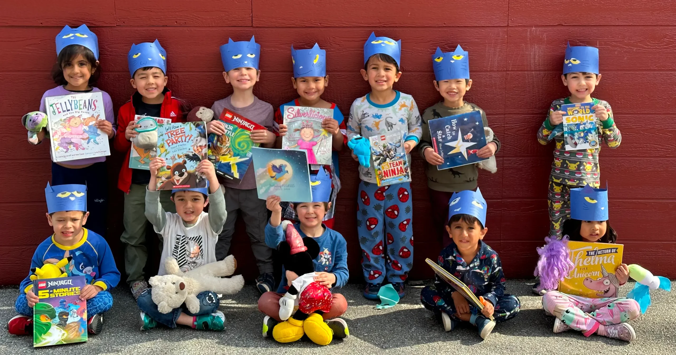 Preschool children at Action Day Schools wearing blue paper hats, holding books, and smiling in front of a red wall.